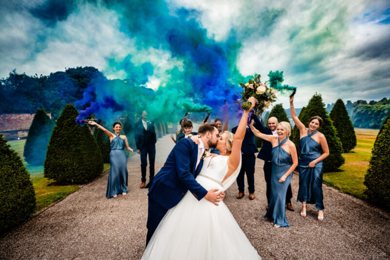 Bride & Groom kissing with wedding party behind them holding blue smoke flares