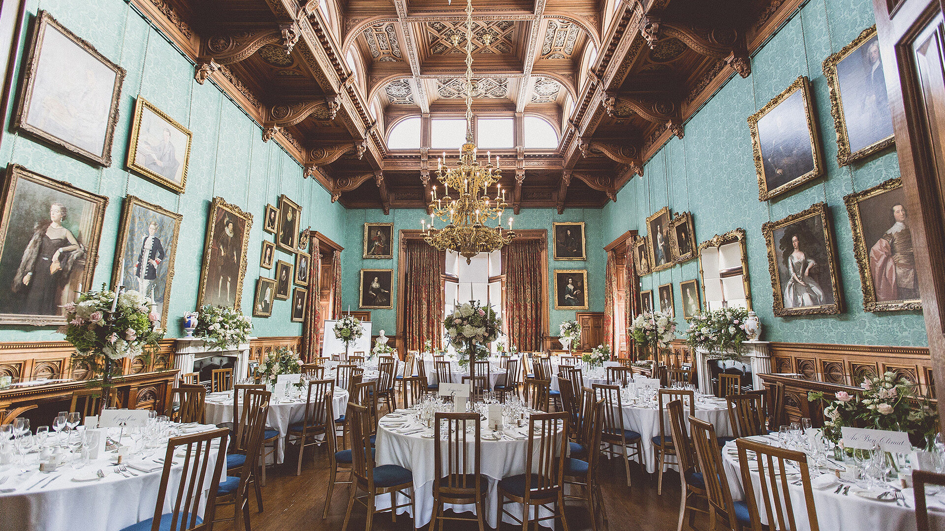 A huge dining room with gold chandelier filled with round tables dressed with white linen, chairs and flowers for a summer wedding