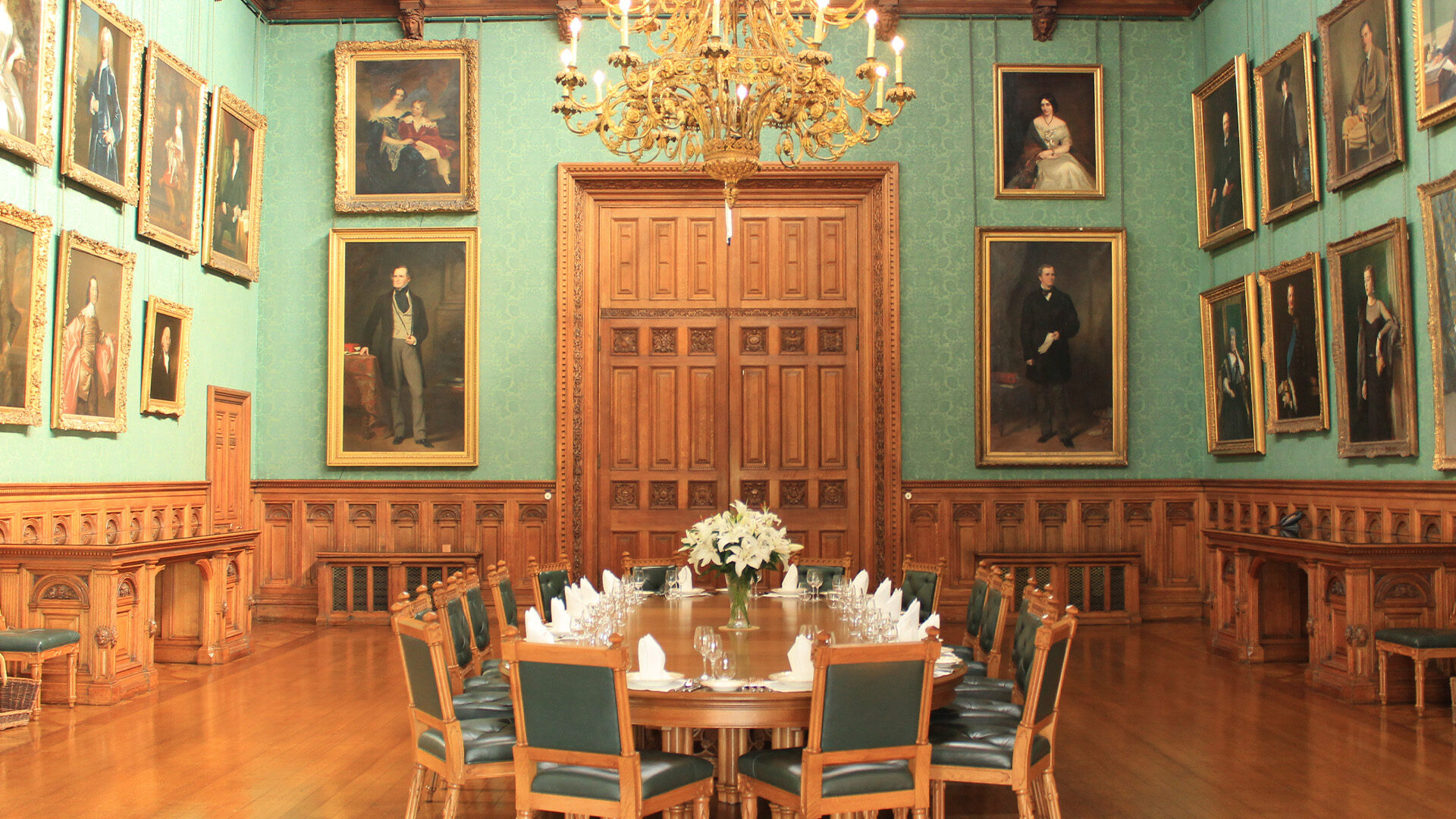 A large dining table surrounded by green leather chairs placed in a huge dining room with paintings hanging on the walls and gold chandelier