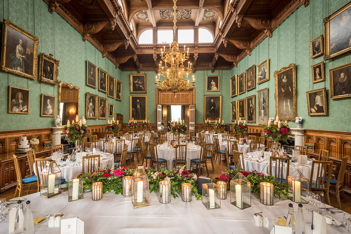 Grand dining room with high ceiling and huge gold chandelier.  Circular tables with blue wooden chairs topped with red bouquets and candles. Paintings lay over a blue wallpaper design.