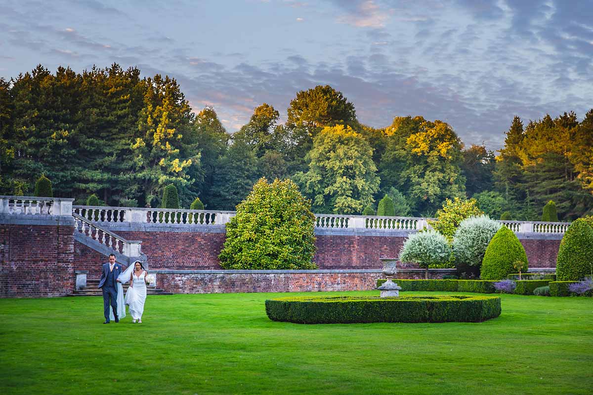 A Bride & Groom walking across huge manicured gardens on their summer wedding day with trees surrounding them.