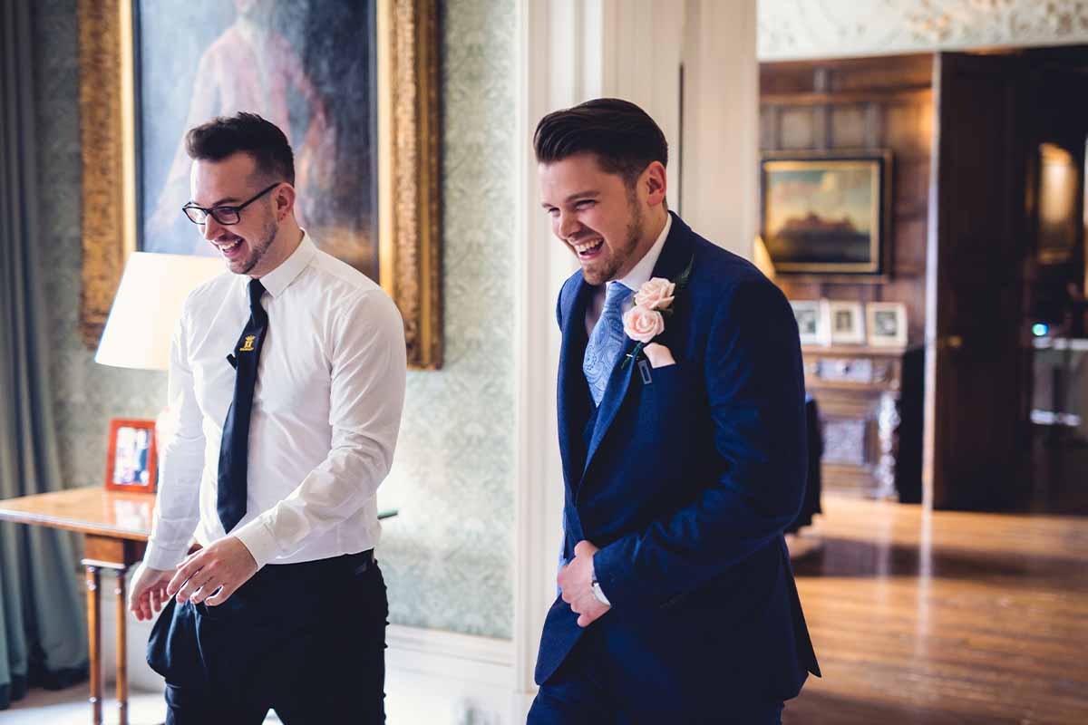 Two men walking together in Knowsley Hall and laughing on wedding day